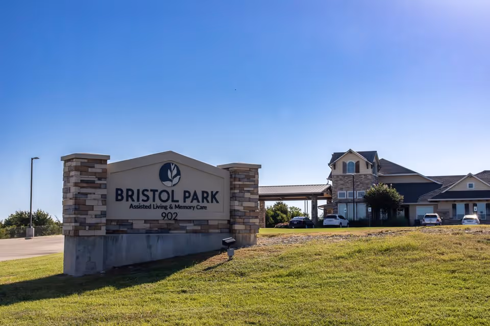 Exterior view of Bristol Park Assisted Living & Memory Care facility with a large stone sign displaying the facility name and address 902, a clear blue sky, and the building with parked cars in the background.