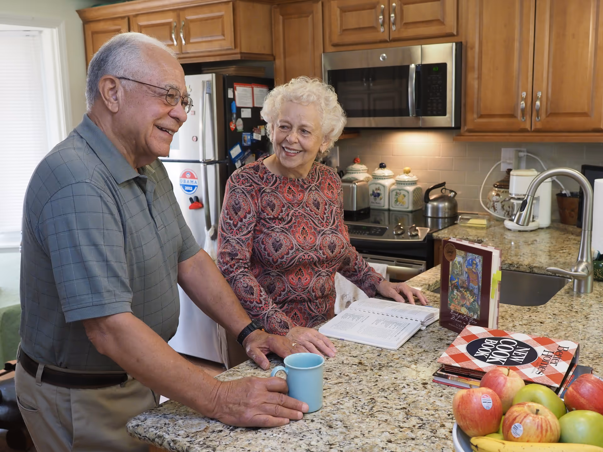 An elderly man and woman standing and smiling together in a kitchen. The man is holding a blue mug while the woman is looking at an open book on the granite countertop. The kitchen has wooden cabinets, a stainless steel microwave, a refrigerator with magnets, and a bowl of fruit on the counter.