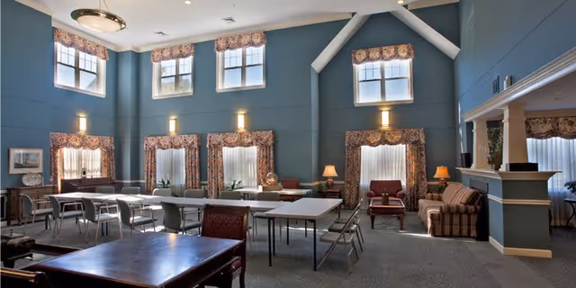 A spacious, well-lit common area with high ceilings and multiple windows with floral curtains. The room contains several tables and chairs arranged for group seating, along with a few armchairs and a sofa near a fireplace. The walls are painted blue, and there are wall-mounted lights providing additional illumination.