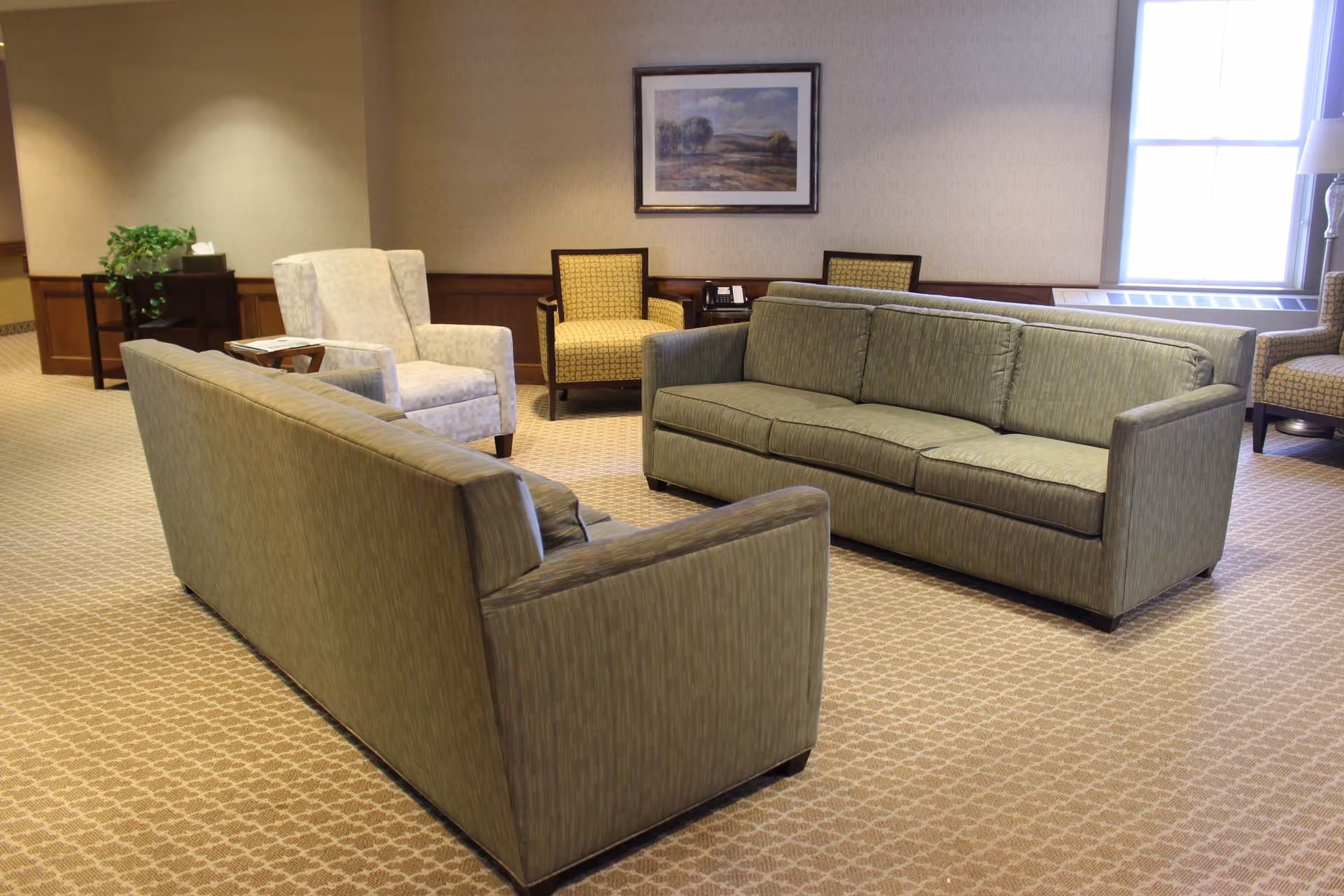 A carpeted sitting area with two green sofas, several armchairs, a framed painting on the wall, and a window letting in light.