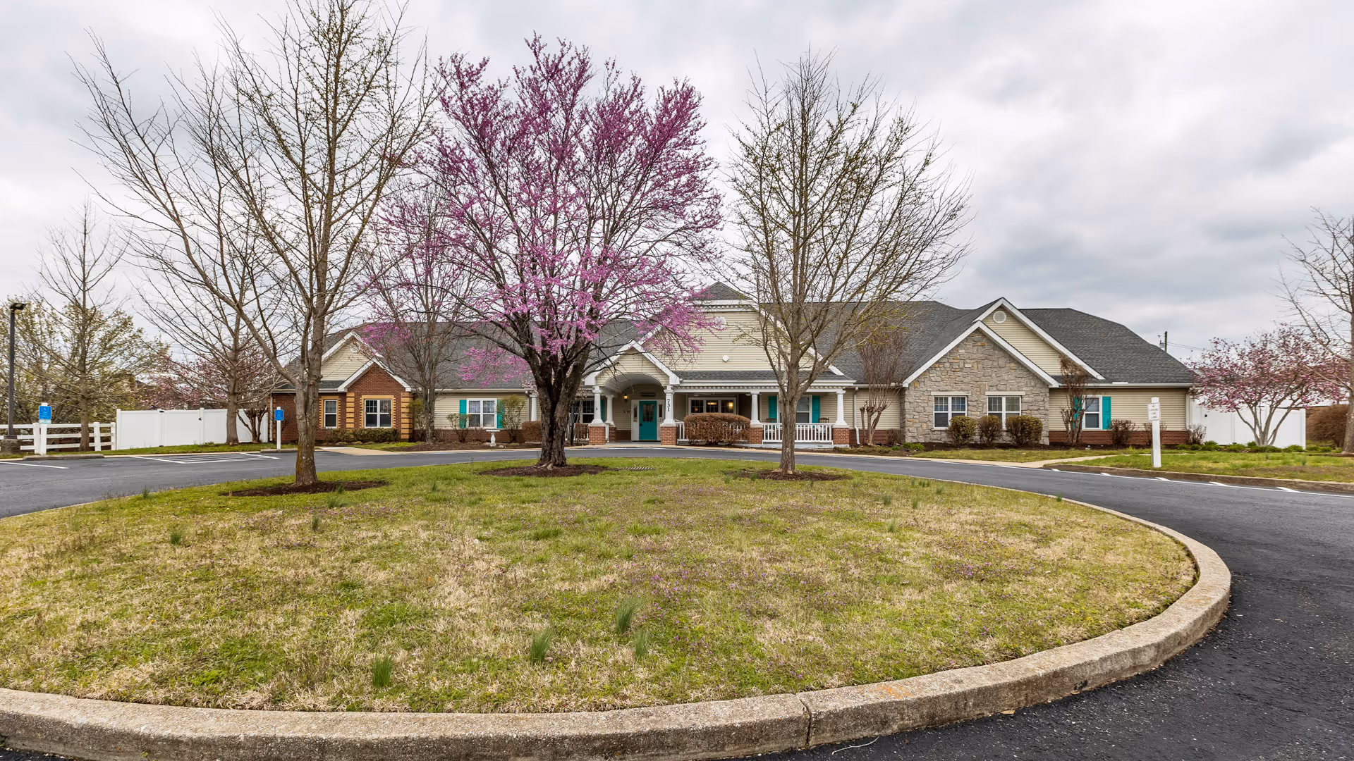 Front exterior view of a single-story senior living facility building with a circular driveway and a grassy roundabout with trees, some of which have pink blossoms, under a cloudy sky.