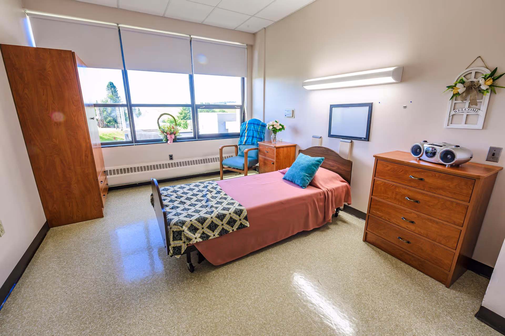 A bright and clean bedroom in a senior living facility with a single bed covered in a pink bedspread and a patterned blanket at the foot. There is a blue pillow on the bed. Next to the bed is a wooden nightstand with a vase of flowers and a wooden chair with a blue blanket draped over it. A large window with white blinds lets in natural light. Across from the bed is a wooden dresser with a radio on top and a decorative wall hanging that says 'Welcome'. The floor is a light-colored linoleum.