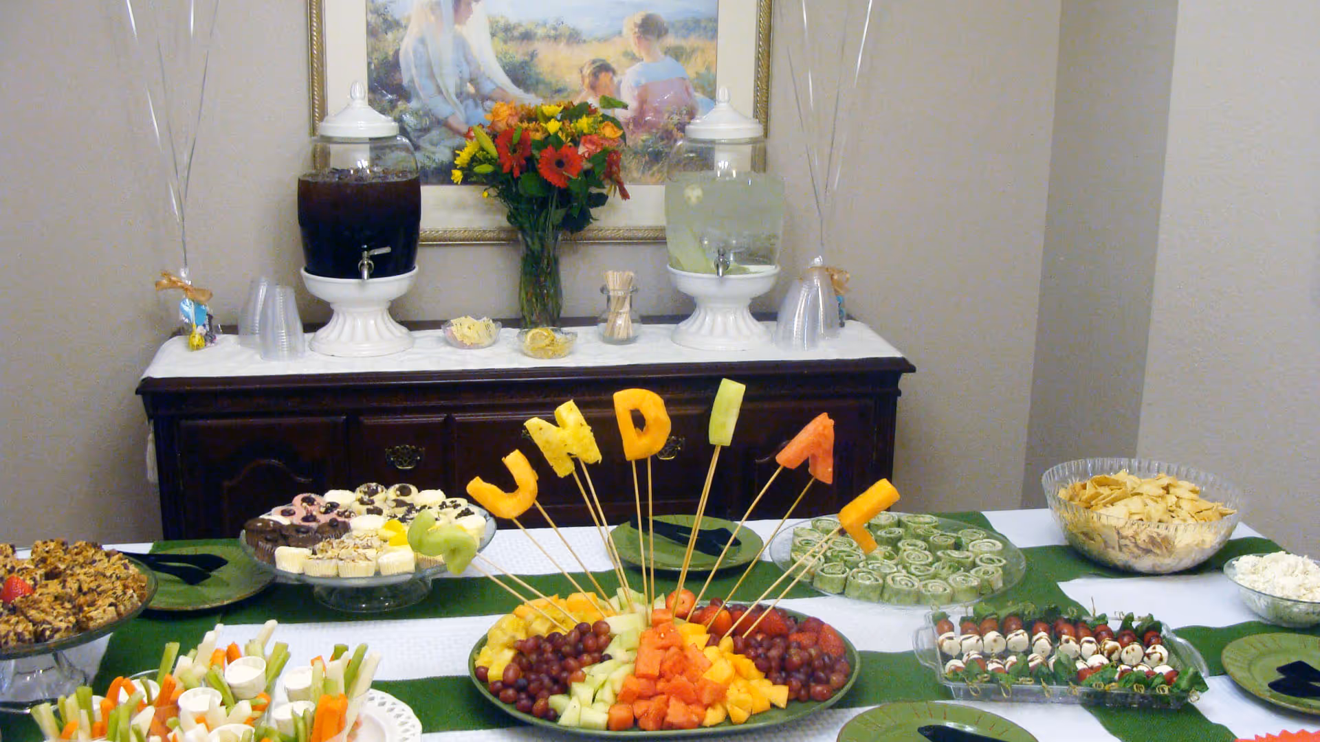 A buffet table set up with a variety of snacks and refreshments including fruit skewers spelling out 'SUNDIAL', vegetable sticks with dip, assorted desserts, crackers, and two large beverage dispensers with dark and light drinks. A vase with colorful flowers and a framed painting are on a wooden sideboard behind the table.