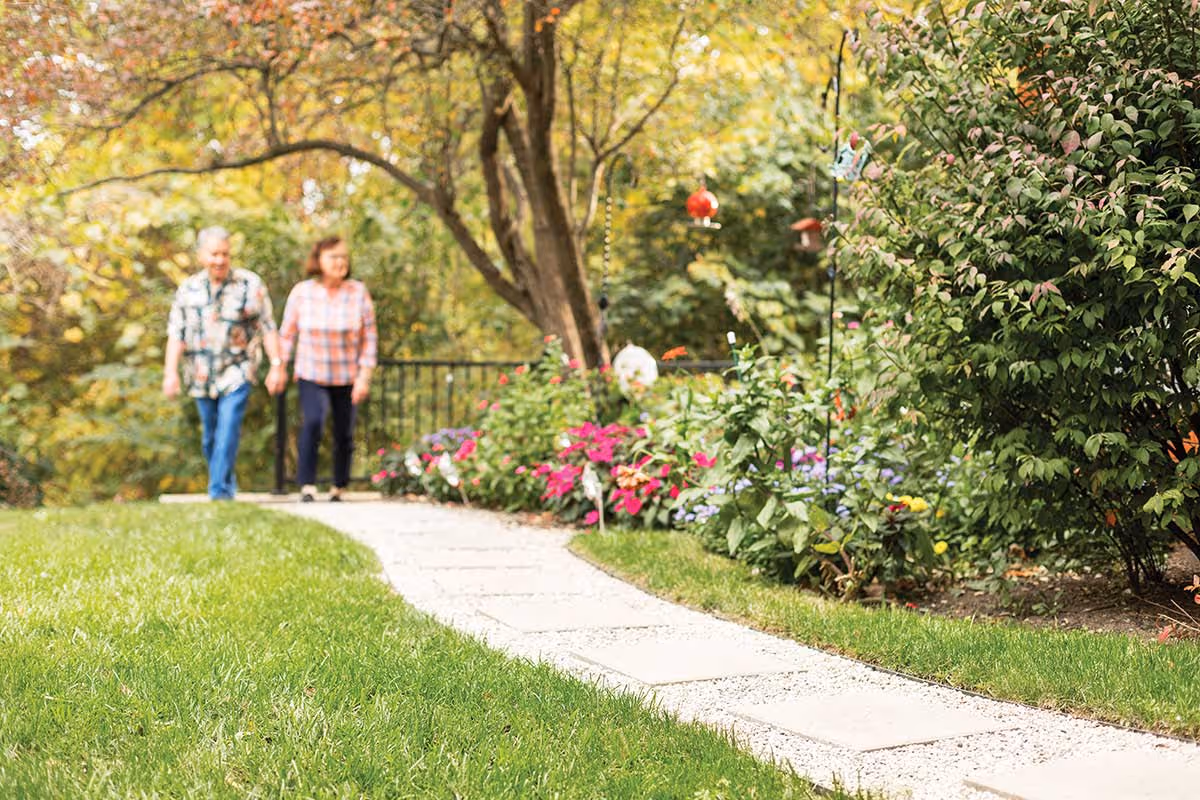 An elderly couple walking hand in hand along a stone pathway in a garden with green grass, colorful flowers, and trees in the background.