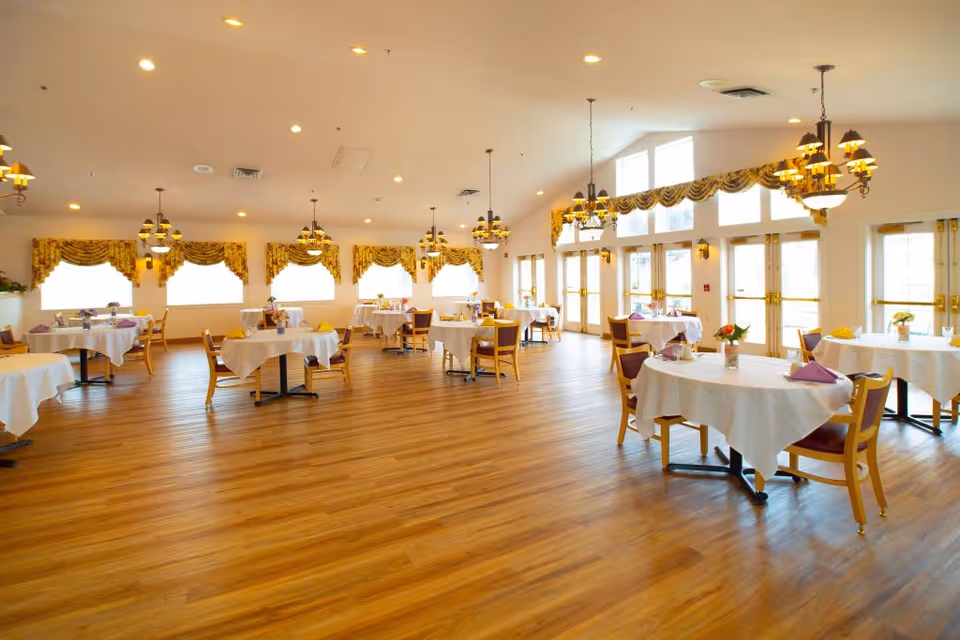 A spacious, well-lit dining room with multiple round tables covered with white tablecloths. Each table has chairs and is set with napkins and small floral centerpieces. Large windows with decorative yellow curtains allow natural light to fill the room, and elegant chandeliers hang from the ceiling. The floor is wooden, and the overall atmosphere is bright and inviting.