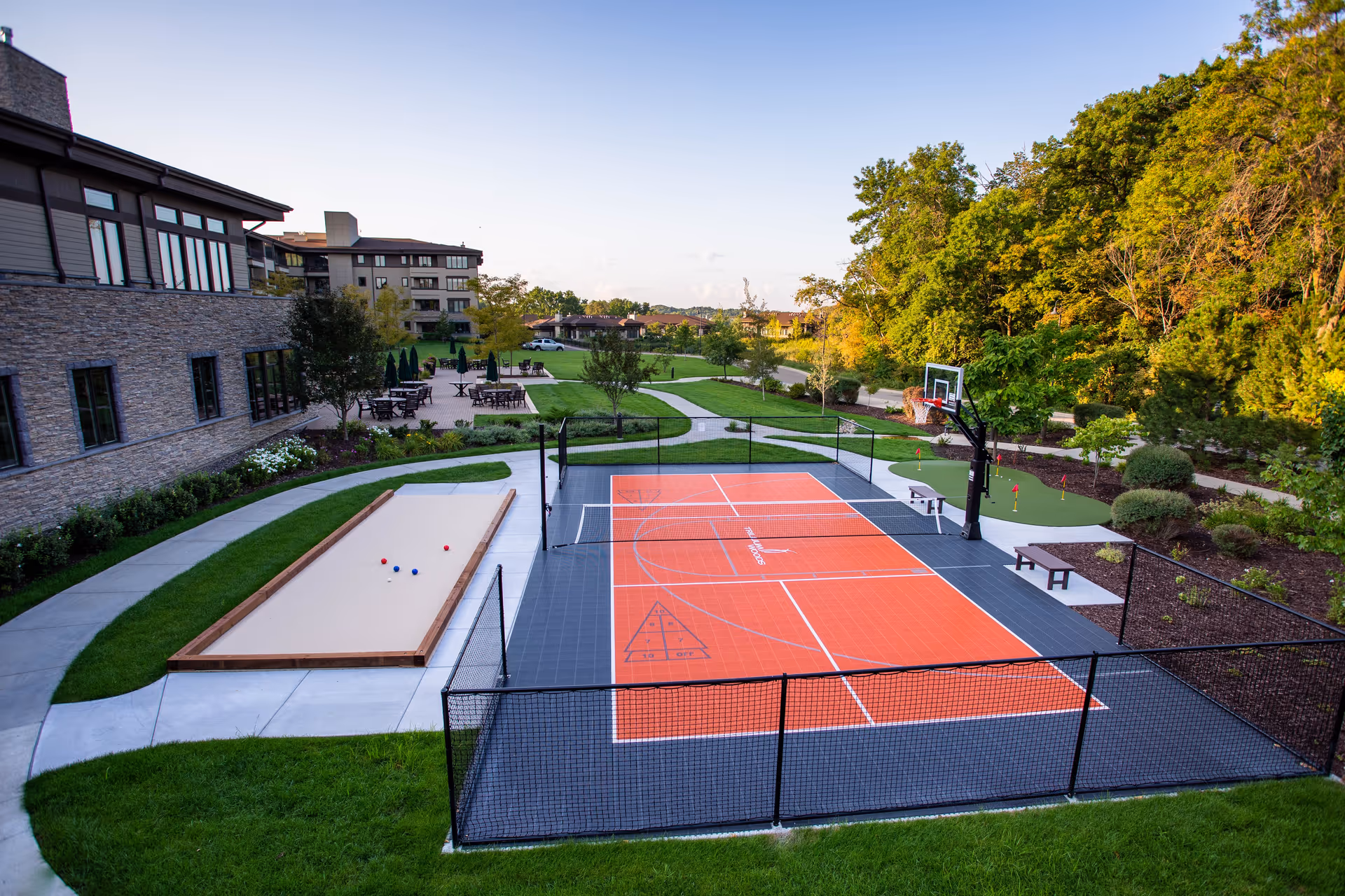 Outdoor recreational area with an orange multi-sport court, a bocce/shuffleboard pit, pathways, seating, and landscaped grounds next to the facility buildings.