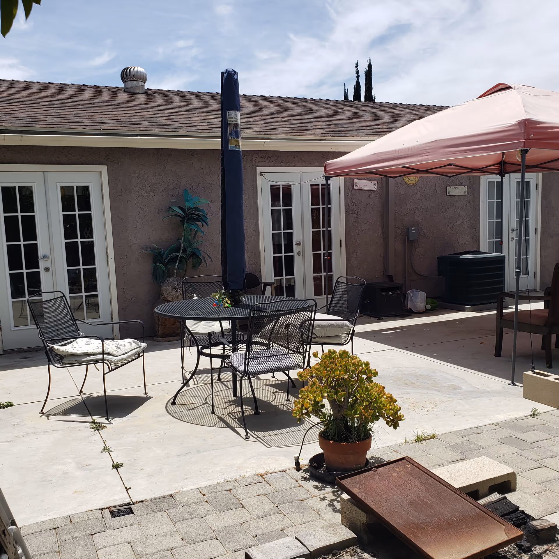 Outdoor patio area with a round metal table and four metal chairs with cushions. A closed umbrella is in the center of the table. There is a potted plant in the foreground and a canopy tent providing shade on the right side. The building wall has three sets of white French doors and some decorative wall hangings.