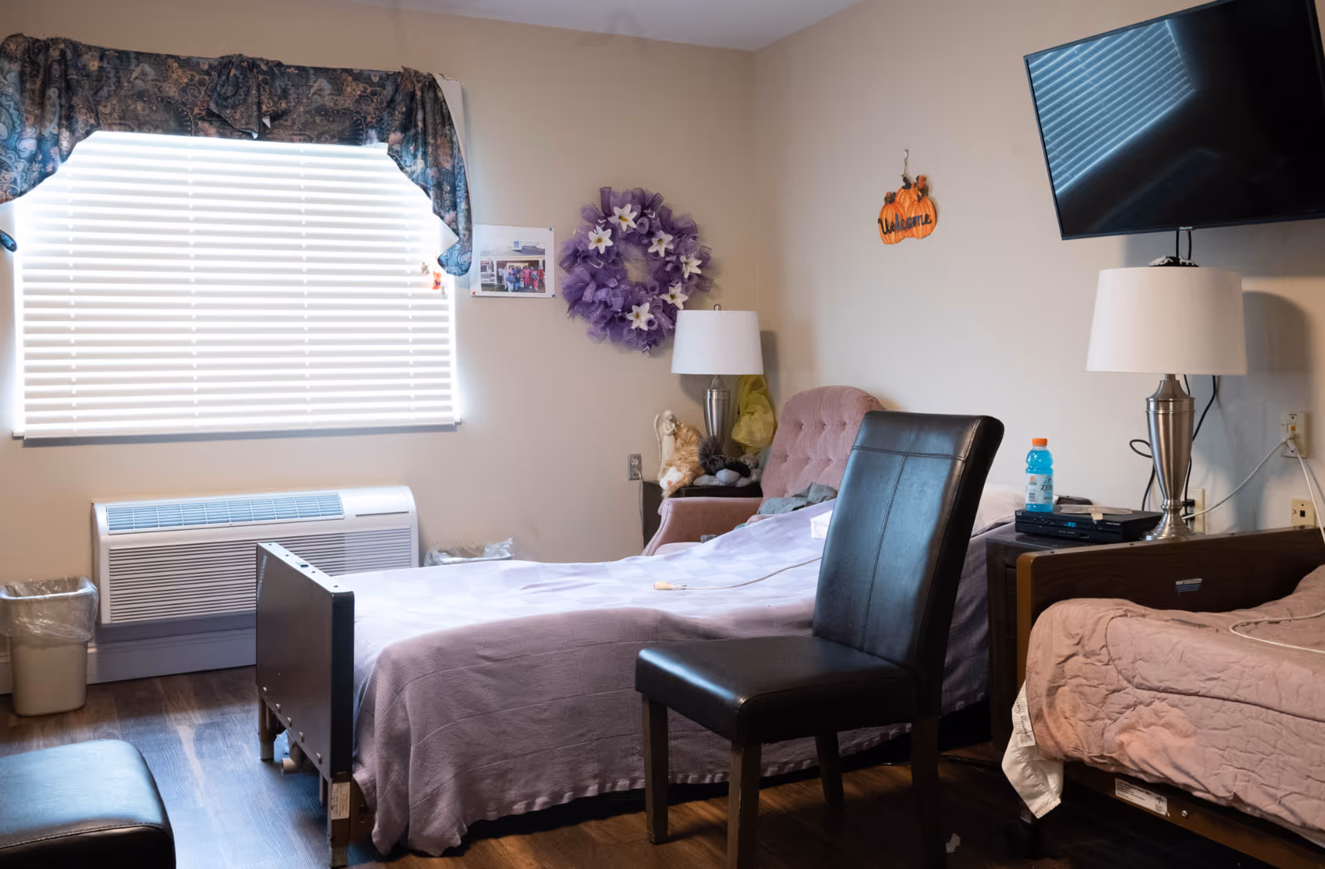 A cozy room with two beds, one covered with a light purple blanket and the other with a pink quilt. A black leather chair is positioned near the bed with the purple blanket. There is a window with closed blinds and a patterned valance. A purple floral wreath and a small photo are hung on the beige wall. A pink armchair with stuffed animals and a silver lamp is in the corner. A flat-screen TV is mounted on the wall above a nightstand with another silver lamp and a blue sports drink bottle. The floor is wooden.