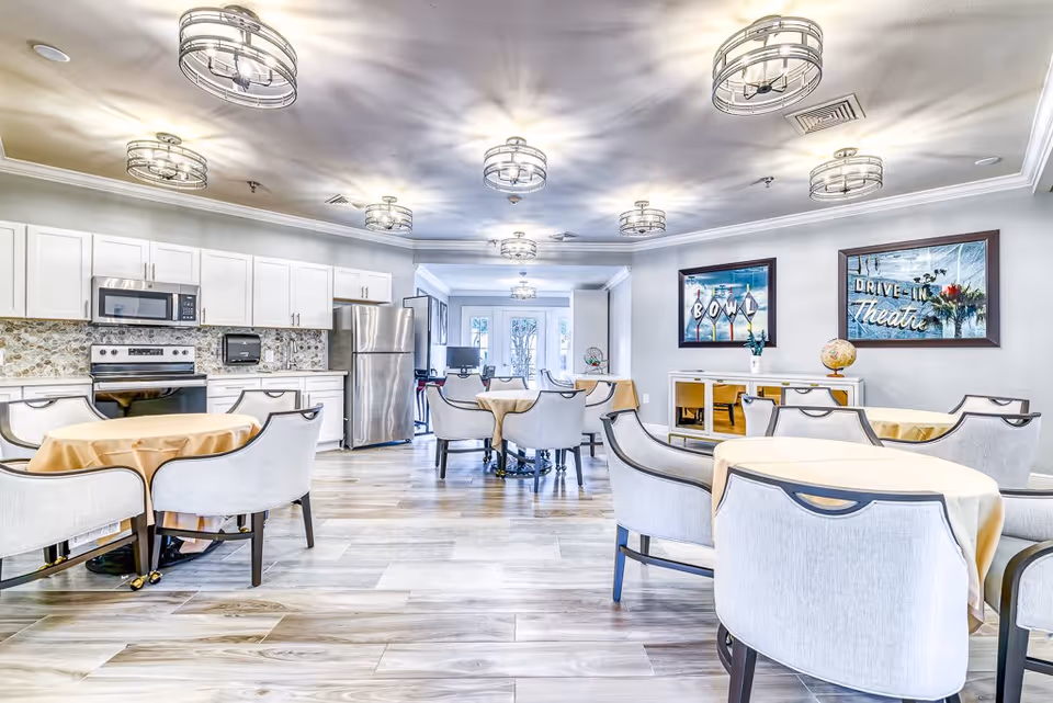 A bright and spacious dining area in a senior living facility featuring multiple round tables covered with beige tablecloths and surrounded by cushioned chairs. The room includes a modern kitchen area with white cabinets, a stainless steel refrigerator, oven, and microwave. The walls are decorated with framed vintage-style posters, and the ceiling has several stylish light fixtures.