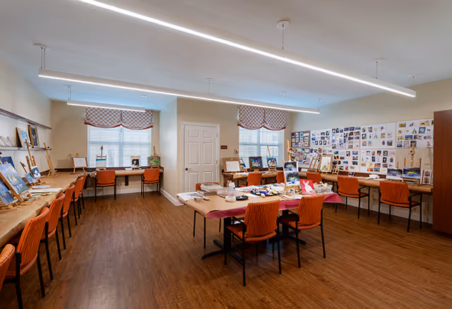 A spacious art room with wooden flooring and multiple orange chairs arranged around tables and along the walls. The tables hold various art supplies and paintings, and the walls are decorated with numerous small artworks and photographs. Two windows with checkered valances allow natural light into the room.