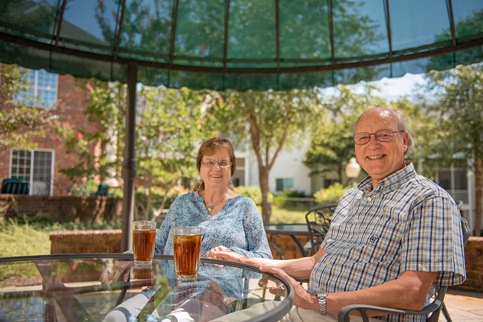 An elderly man and woman sitting at a glass outdoor table under a green canopy, smiling and enjoying iced tea on a sunny day with trees and buildings in the background.