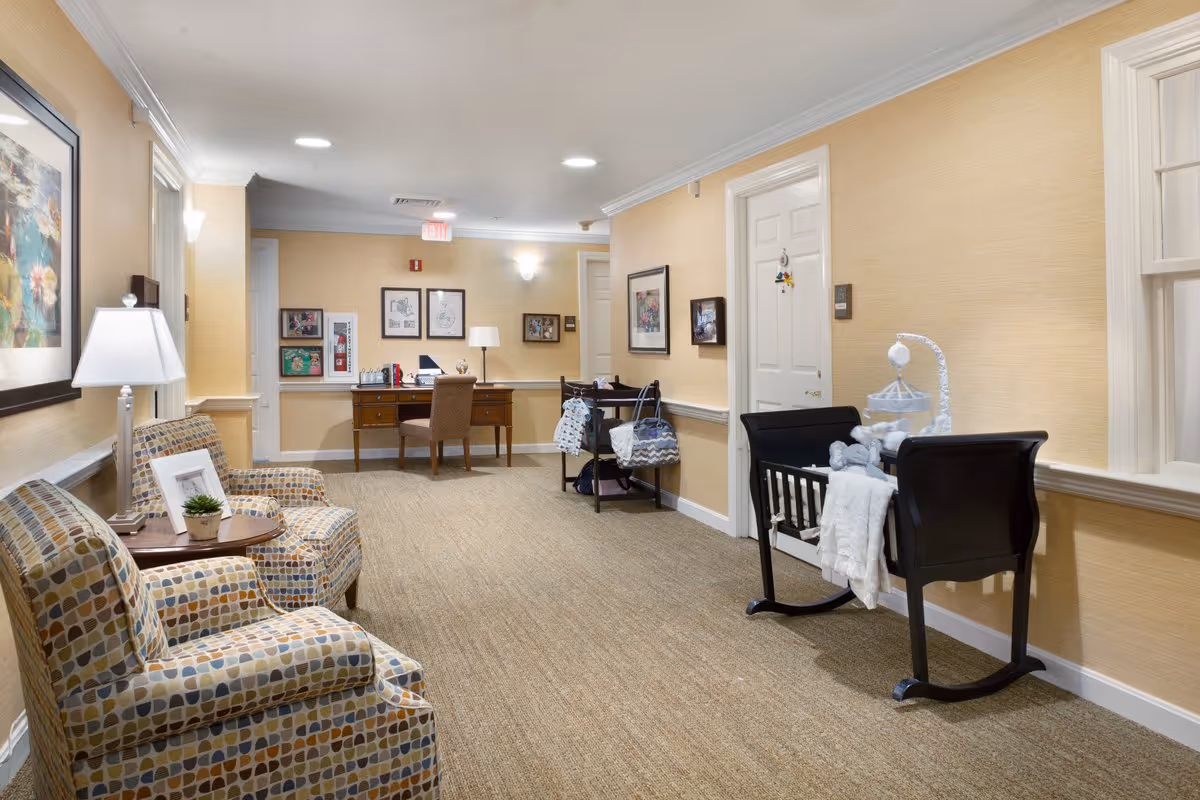 A well-lit hallway in a senior living facility with beige walls and carpeted floor. On the left side, there are two patterned armchairs with a small round table and a lamp between them. On the right side, there is a black wooden cradle with a baby mobile and blankets. Further down the hallway, there is a wooden desk with a chair, a lamp, and some framed pictures on the wall behind it. Several framed artworks and photographs decorate the walls.