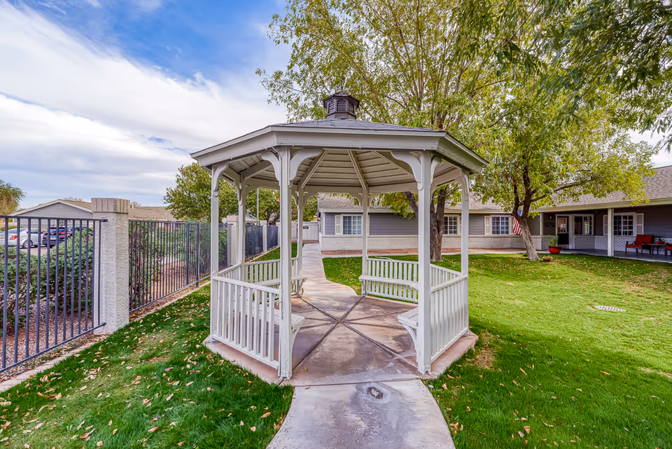 A white wooden gazebo with benches inside situated on a concrete path in a grassy courtyard area. Surrounding the gazebo are trees with green leaves and a building with gray siding and white trim in the background. A black metal fence and parked cars are visible on the left side under a partly cloudy sky.