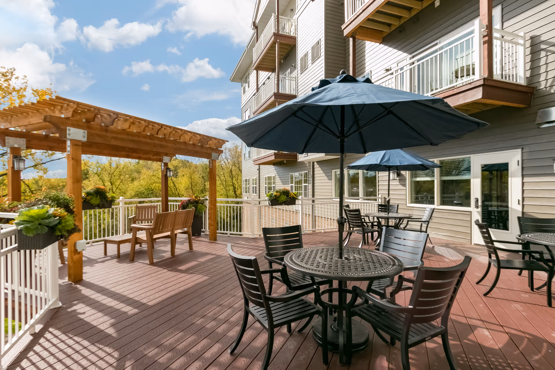 Outdoor patio area at a senior living facility with round metal tables and chairs under large blue umbrellas, wooden benches under a pergola, potted plants hanging on the railing, and a multi-story building with balconies in the background under a partly cloudy sky.