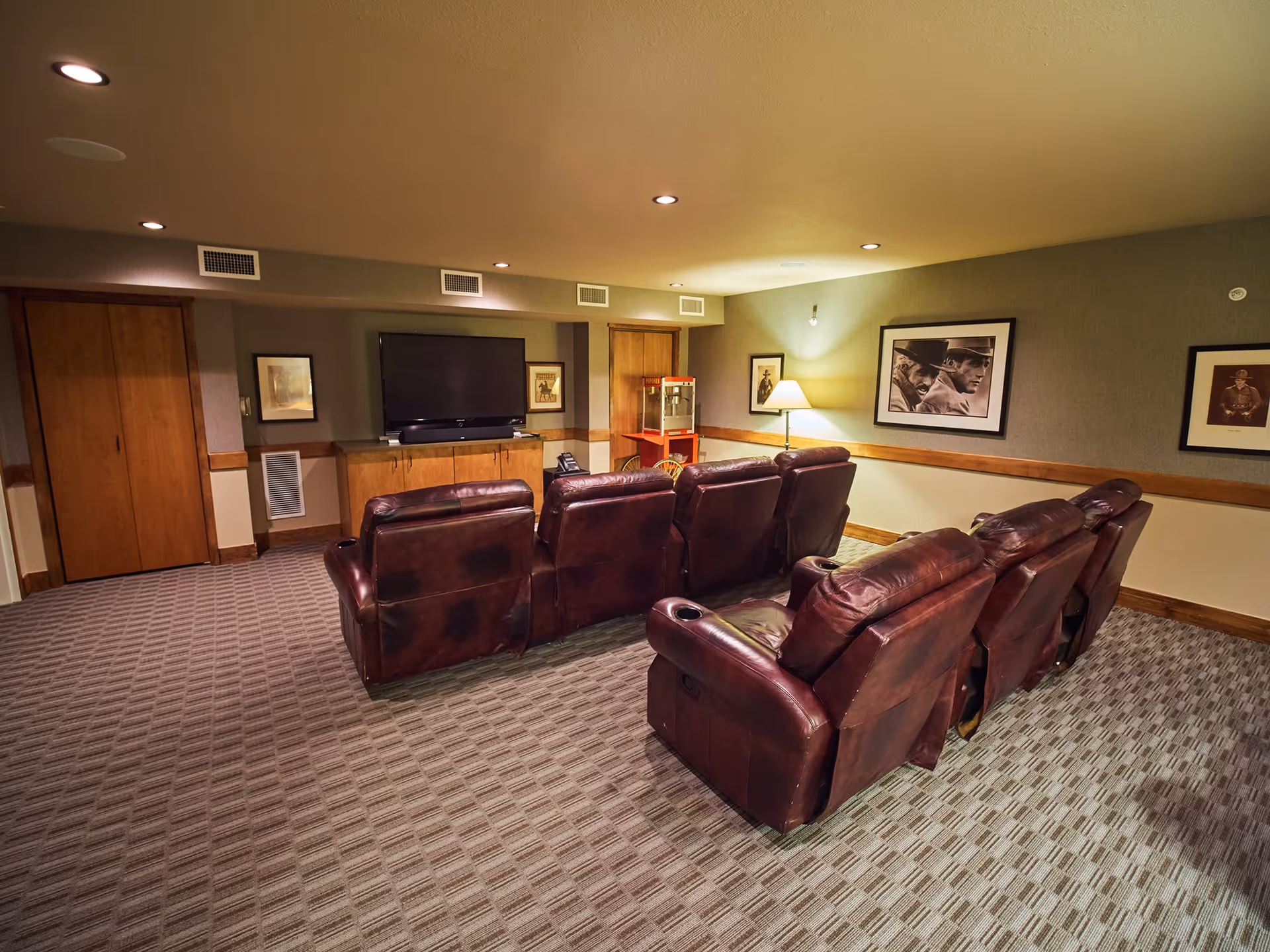 A cozy media room with two rows of brown leather recliner chairs facing a large flat-screen TV mounted on a wooden cabinet. The room has carpeted flooring, recessed ceiling lights, framed black and white photographs on the walls, a floor lamp, and a popcorn machine in the corner.