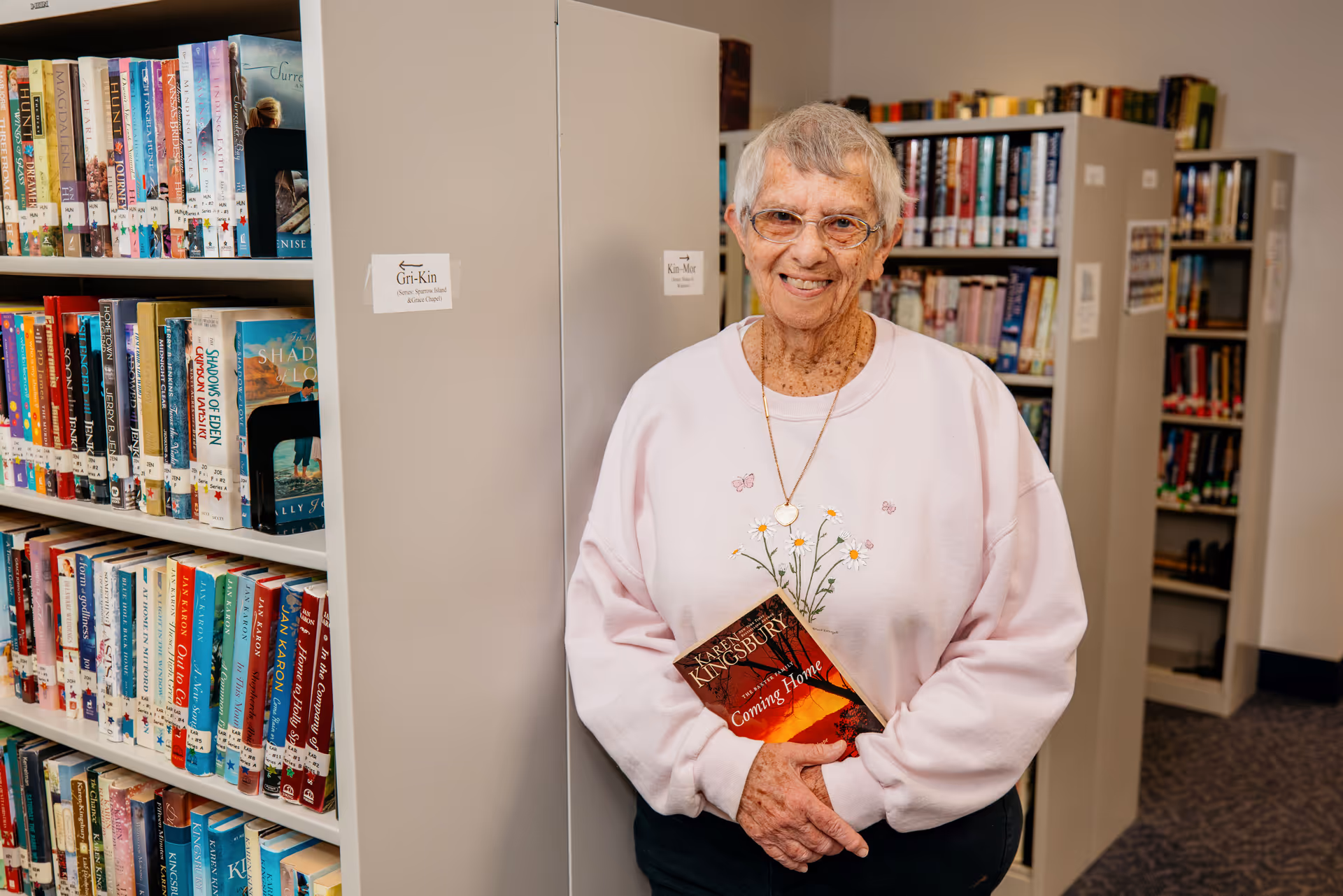 An elderly woman with short gray hair and glasses stands smiling in a library area of a nursing home, holding a book titled 'Coming Home' by Karen Kingsbury. She is wearing a light pink sweatshirt with a floral design and a heart-shaped necklace. Behind her are shelves filled with books.