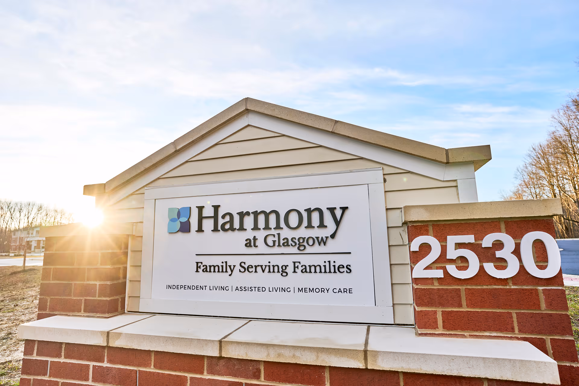 Outdoor entrance sign reading "Harmony at Glasgow Family Serving Families" on a brick monument with the street number 2530 at sunrise.