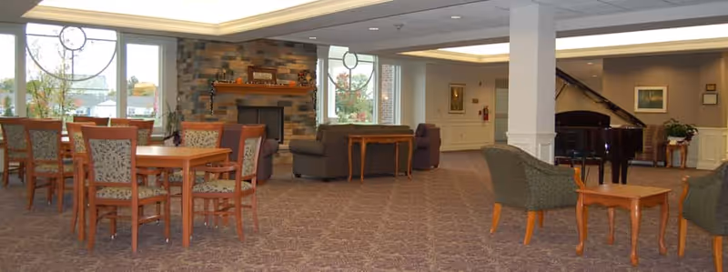 A spacious assisted living common area featuring a carpeted floor, wooden tables with chairs, a stone fireplace, a grand piano, and comfortable armchairs arranged around small tables. Large windows allow natural light to fill the room.