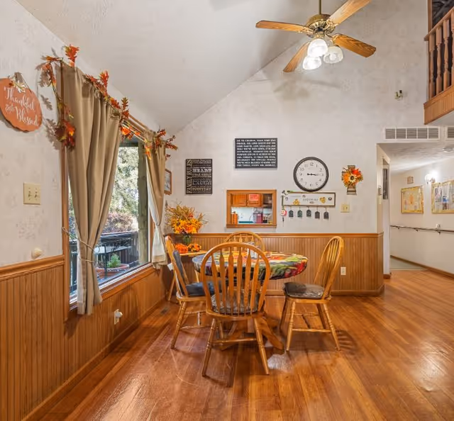 A cozy dining area in a senior living facility with a round wooden table covered with a colorful tablecloth and four wooden chairs. The room has wood-paneled walls and hardwood floors. A large window with beige curtains decorated with autumn leaves lets in natural light. The wall features a clock, decorative signs, and a small pass-through window to the kitchen. A ceiling fan with lights hangs above the table.