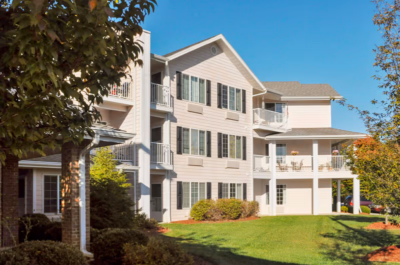Three-story light-colored apartment building with balconies, many windows, and a manicured lawn under a clear blue sky.