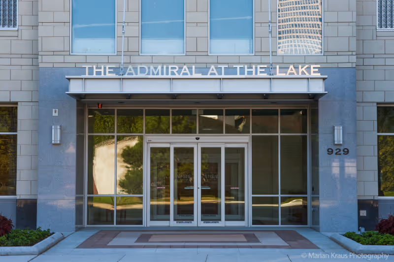 Front entrance of The Admiral at the Lake building with glass doors and large windows, flanked by stone walls and greenery on either side. The building number 929 is visible on the right side.