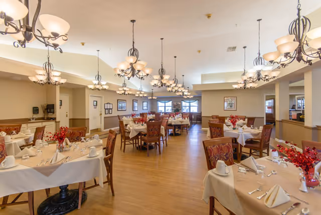 A spacious dining room with multiple round tables covered with beige tablecloths, each set with white napkins, cups, and silverware. The tables have red floral centerpieces. The room features wooden flooring, beige walls with wainscoting, and multiple ornate chandeliers hanging from the ceiling. Windows are visible at the far end of the room, allowing natural light to enter.