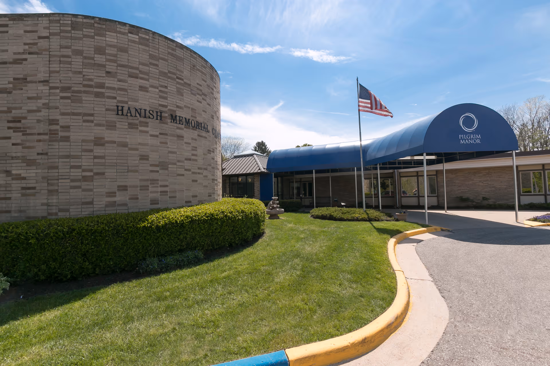 Exterior view of a building with a curved brick wall labeled 'HANISH MEMORIAL' and a blue canopy entrance with the logo and name 'PILGRIM MANOR'. An American flag is flying on a flagpole near the entrance, and there is a well-maintained lawn with bushes and a paved driveway.
