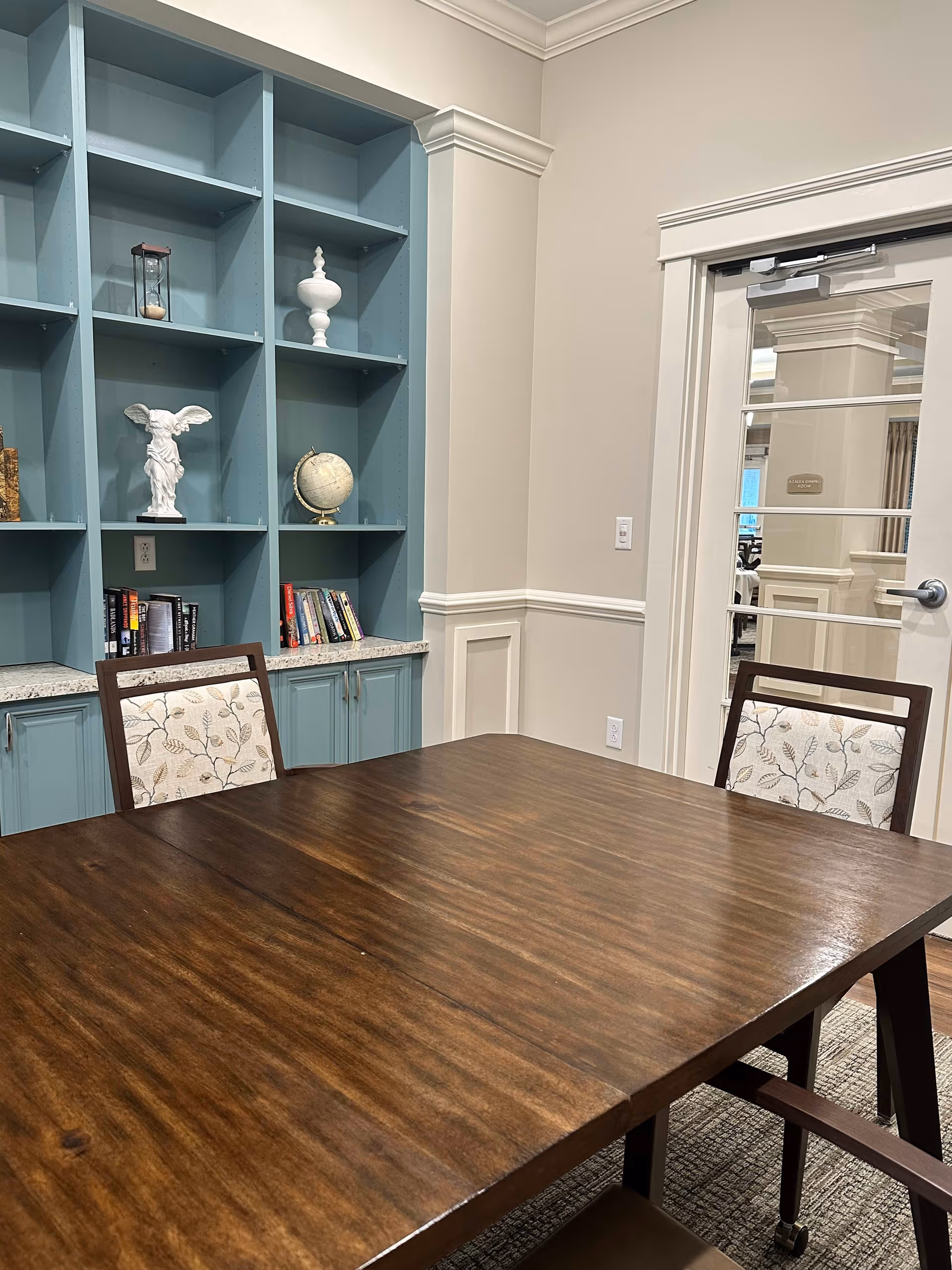 A room with a wooden table and two chairs with floral upholstery. Behind the table is a built-in blue bookshelf with various decorative items including a small globe, a white statue, an hourglass, and several books. To the right, there is a white door with glass panels leading to another room.