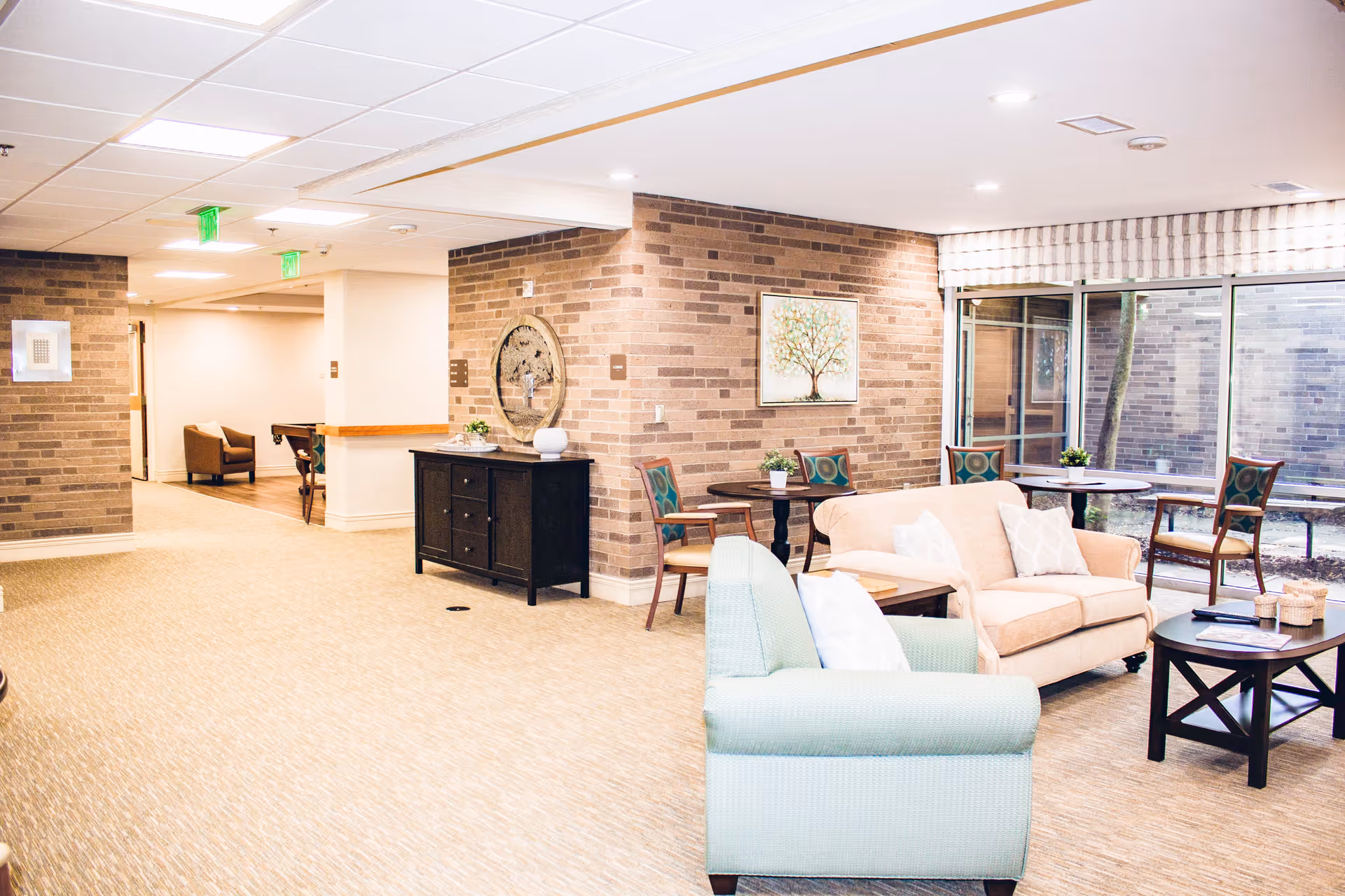 A bright and spacious senior living facility common area with beige carpet, brick accent walls, and a white ceiling with recessed lighting. The room features comfortable seating including a light green armchair and a beige sofa with white pillows, several wooden tables with chairs, and decorative elements such as a round mirror, framed artwork, and small potted plants. Large windows provide natural light and a view of an outdoor courtyard.