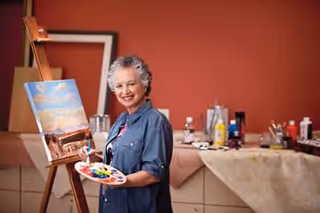 An elderly woman smiling while holding a paint palette and brush, standing in front of an easel with a painting in progress in a room with art supplies and a reddish-brown wall.