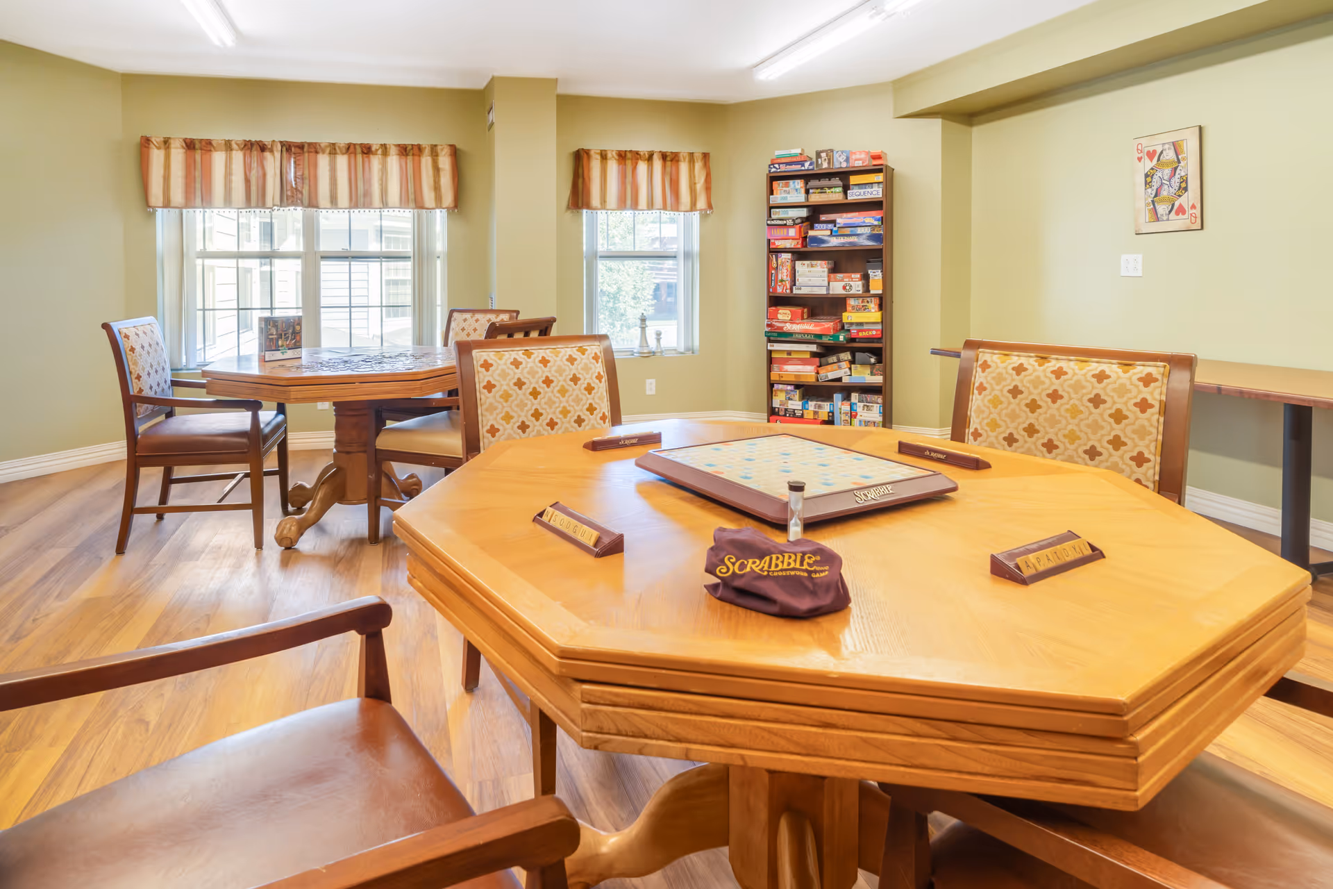 Bright activity room with wooden game tables and chairs, a Scrabble board on the table and a shelf of board games.