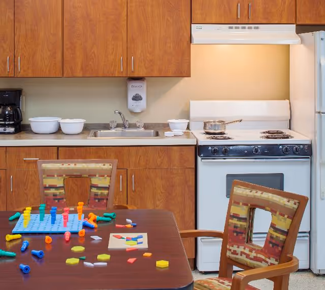 A kitchen area with wooden cabinets, a white stove with a pot on it, a white refrigerator, and a sink. In the foreground, there is a table with colorful pegboard toys and two chairs with patterned upholstery.