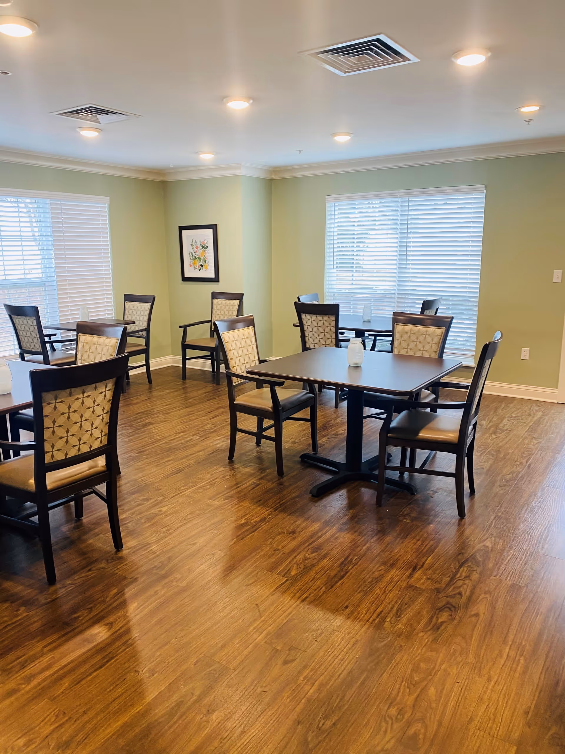 A dining room with several square tables and cushioned chairs arranged neatly on a wooden floor. The walls are painted light green with two large windows covered by white blinds letting in natural light. A framed floral artwork is hung on one wall.