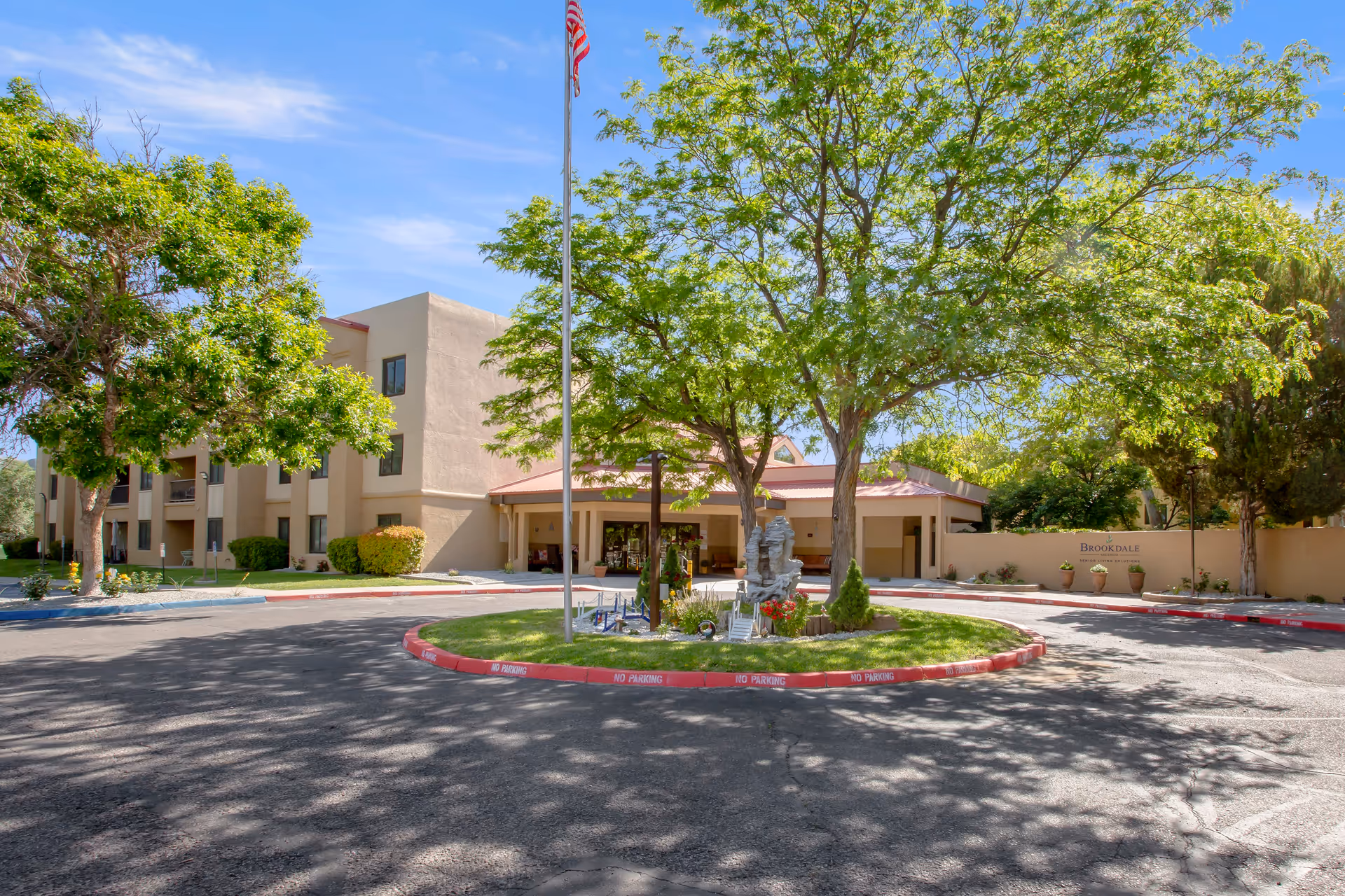 Exterior view of Brookdale Valencia senior living facility showing a circular driveway with a landscaped island featuring trees, a flagpole with an American flag, and a decorative sculpture. The building is beige with multiple windows and a covered entrance. The sky is clear and blue.