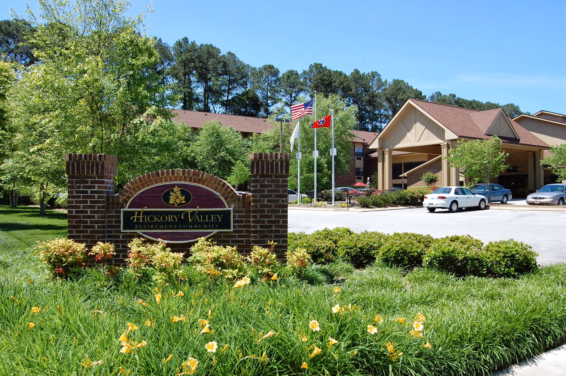Front entrance of Hickory Valley senior living community showing a brick sign, landscaped flowers, flags, parked cars, and the building facade.