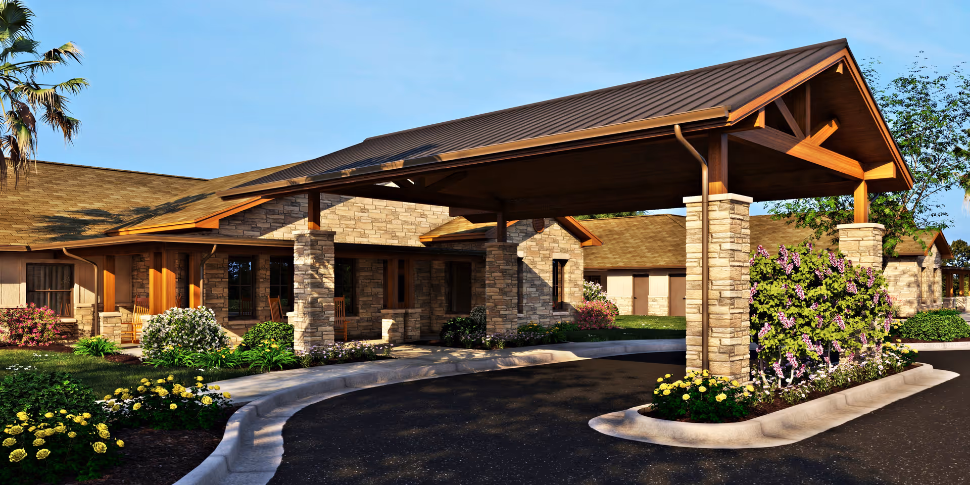 Exterior view of a senior living facility with a covered driveway entrance supported by stone pillars. The building features stone and wood accents, surrounded by landscaped greenery and flowering plants under a clear blue sky.