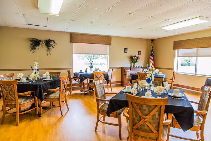 A bright dining room with several wooden tables covered with black tablecloths, each set with white napkins, cups, and plates. Wooden chairs with patterned cushions surround the tables. The room has large windows with beige blinds, a wooden floor, and an American flag in the corner. Floral decorations are placed on the tables and on a small wooden cabinet against the wall.