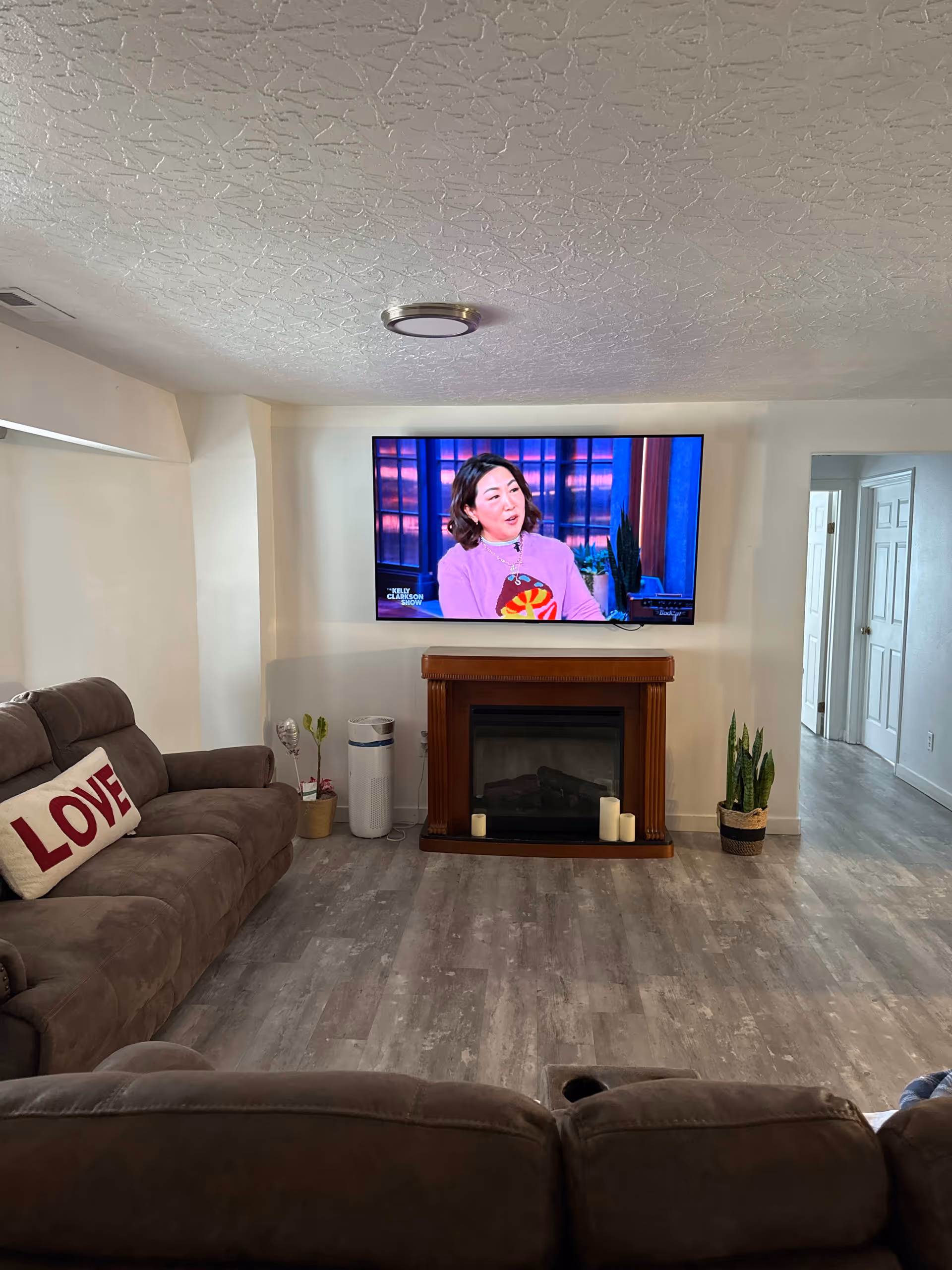 A cozy living room with a large flat-screen TV mounted on the wall above a wooden electric fireplace. The TV is showing a woman in a purple top speaking on a talk show. There are two brown reclining sofas, one with a white pillow that has the word 'LOVE' in red letters. The room has light-colored walls, a textured ceiling, and gray wood-look flooring. There are a few potted plants and candles near the fireplace, and a hallway with white doors is visible to the right.