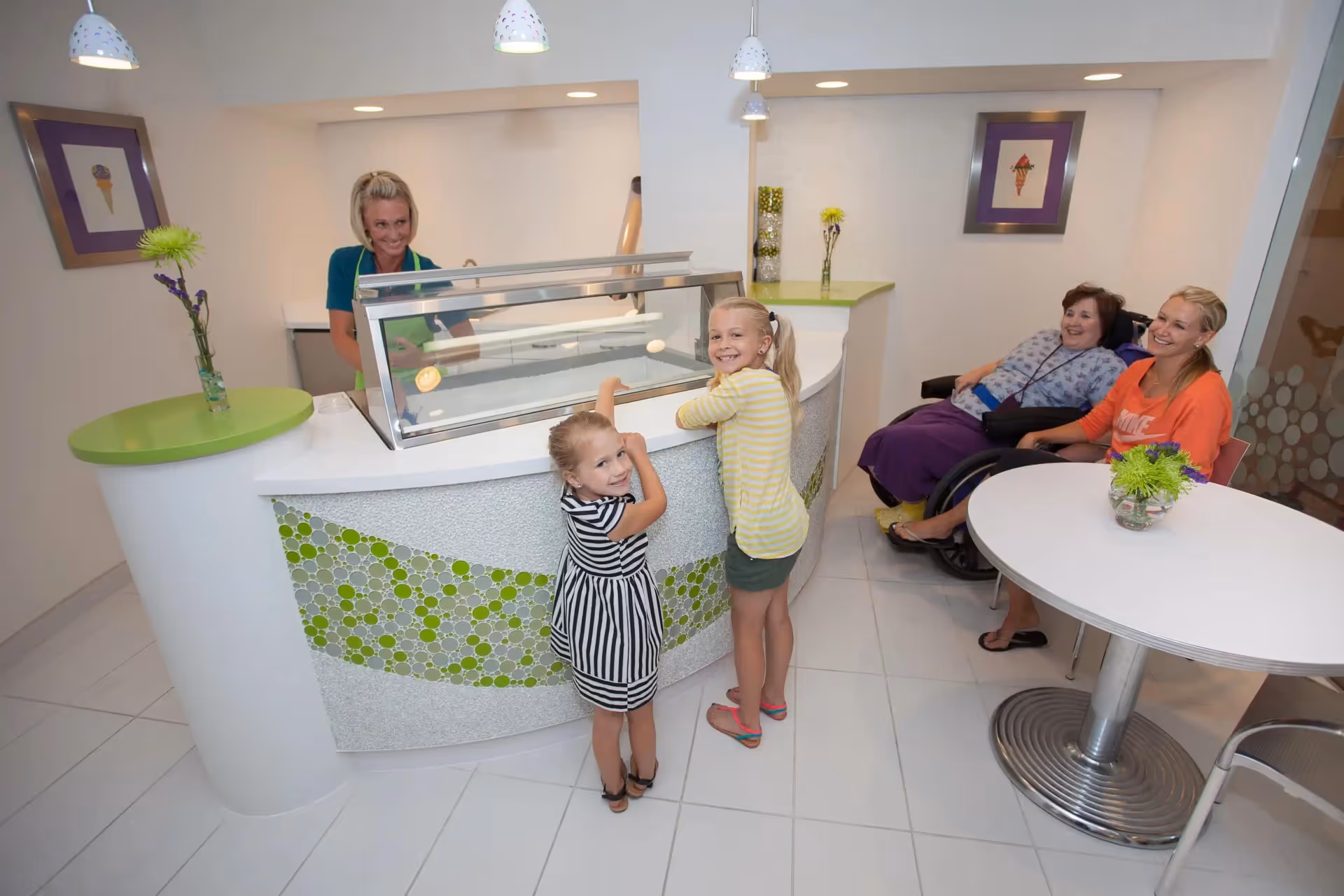 Two young girls stand at a curved service counter with a staff member behind the glass display while two women, one in a wheelchair, sit at a nearby table in a bright dining area.