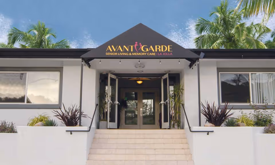 Front exterior view of Avantgarde Senior Living & Memory Care facility in La Jolla with steps leading up to double glass doors under a black awning with the facility name and logo, surrounded by plants and palm trees.
