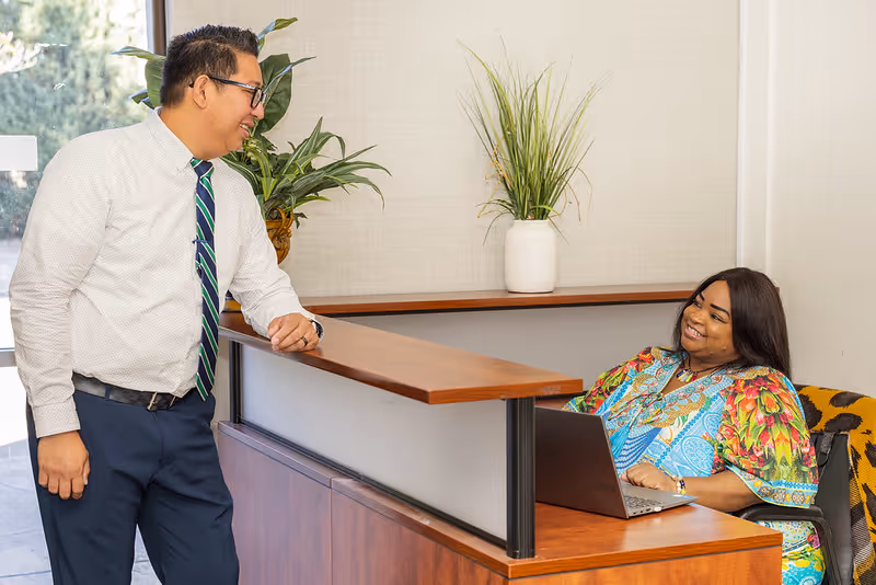 A man in business attire with glasses and a striped tie is standing and leaning on a wooden reception desk, smiling and talking to a woman seated behind the desk. The woman is wearing a colorful patterned dress and is smiling back while using a laptop. There are two potted plants on the desk and a window in the background.