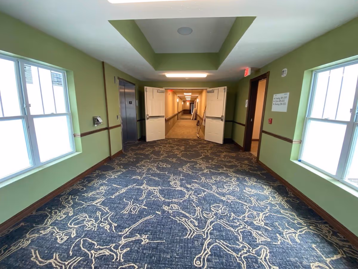 Interior hallway of a senior living facility with patterned carpet, green walls, windows, an elevator, and double doors opening to a long corridor.
