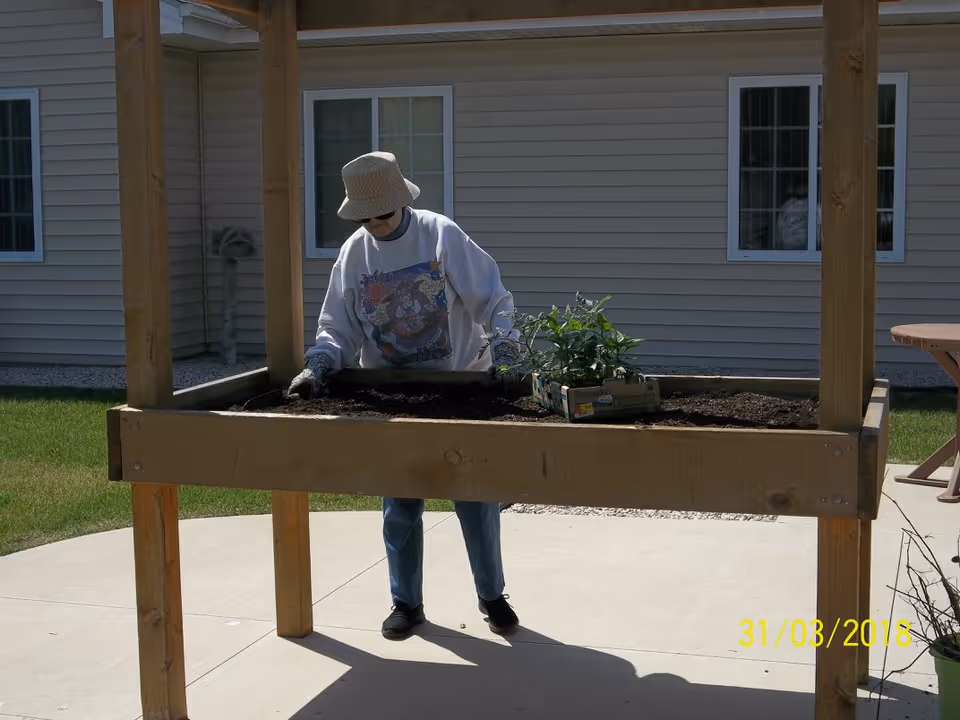 An elderly person wearing a hat, sunglasses, gloves, and a sweatshirt is gardening at a raised wooden planter filled with soil and a small plant. The scene is outdoors on a concrete patio with a beige building with windows in the background.