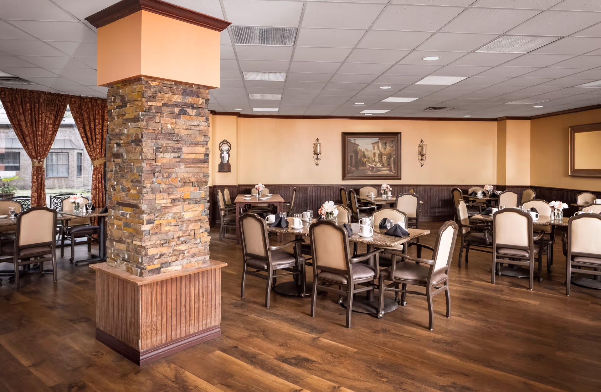 A dining room in an assisted living facility with multiple tables and chairs arranged neatly. Each table is set with white mugs, black napkins, and small floral centerpieces. The room features a stone pillar in the center, wood flooring, and warm beige walls adorned with framed artwork and sconces. Large windows with patterned curtains allow natural light to enter.
