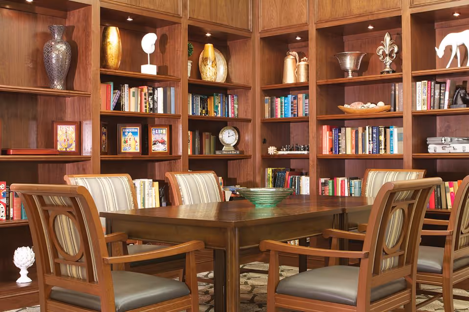 A cozy library or reading room with wooden bookshelves filled with books and decorative items. In the center, there is a wooden table surrounded by six wooden chairs with striped cushions. The room has warm lighting and a comfortable, inviting atmosphere.