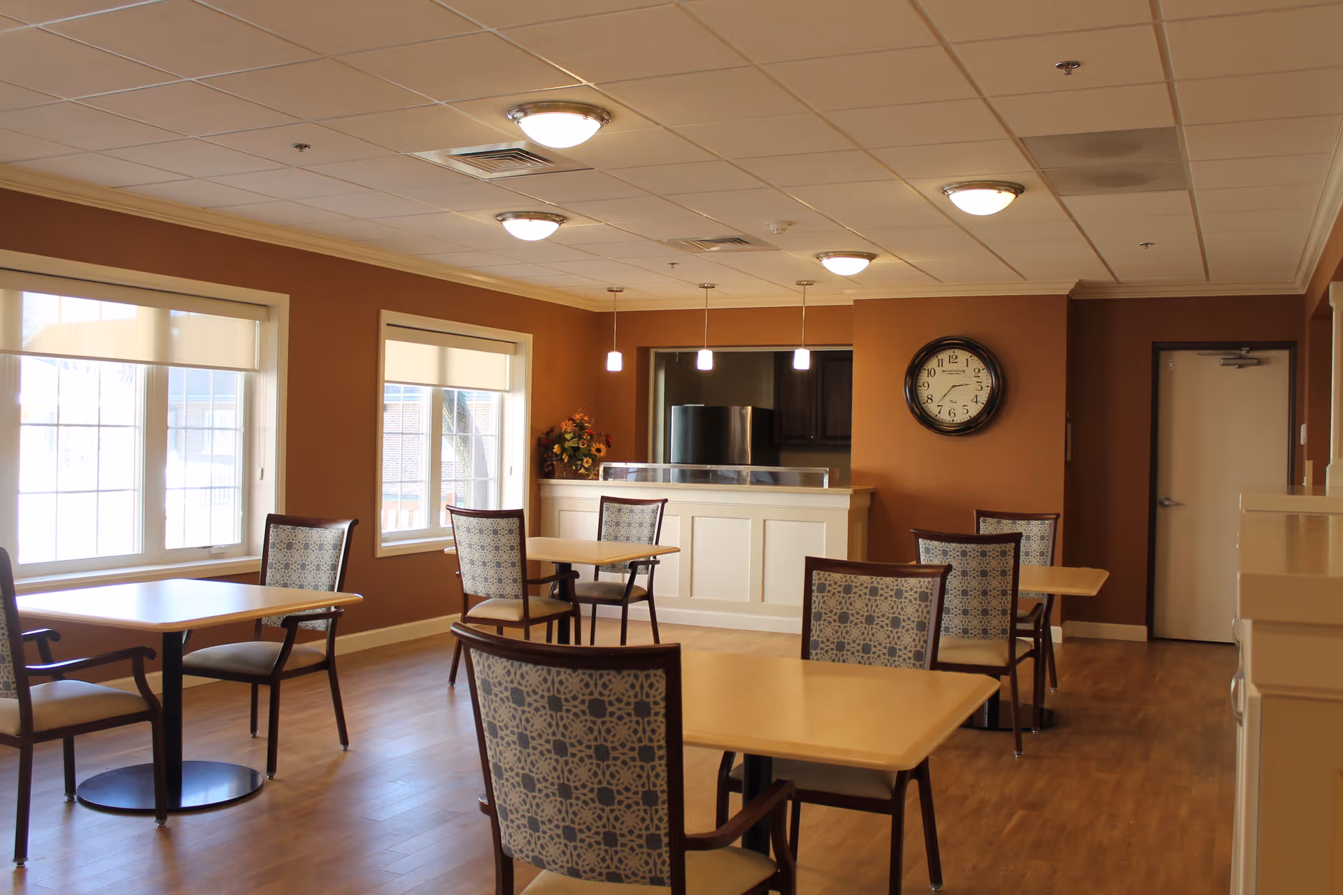 A dining room with several wooden tables and patterned chairs arranged neatly. The room has large windows with white blinds, warm brown walls, and a wooden floor. There is a clock on the wall and a counter area with pendant lights hanging above it.