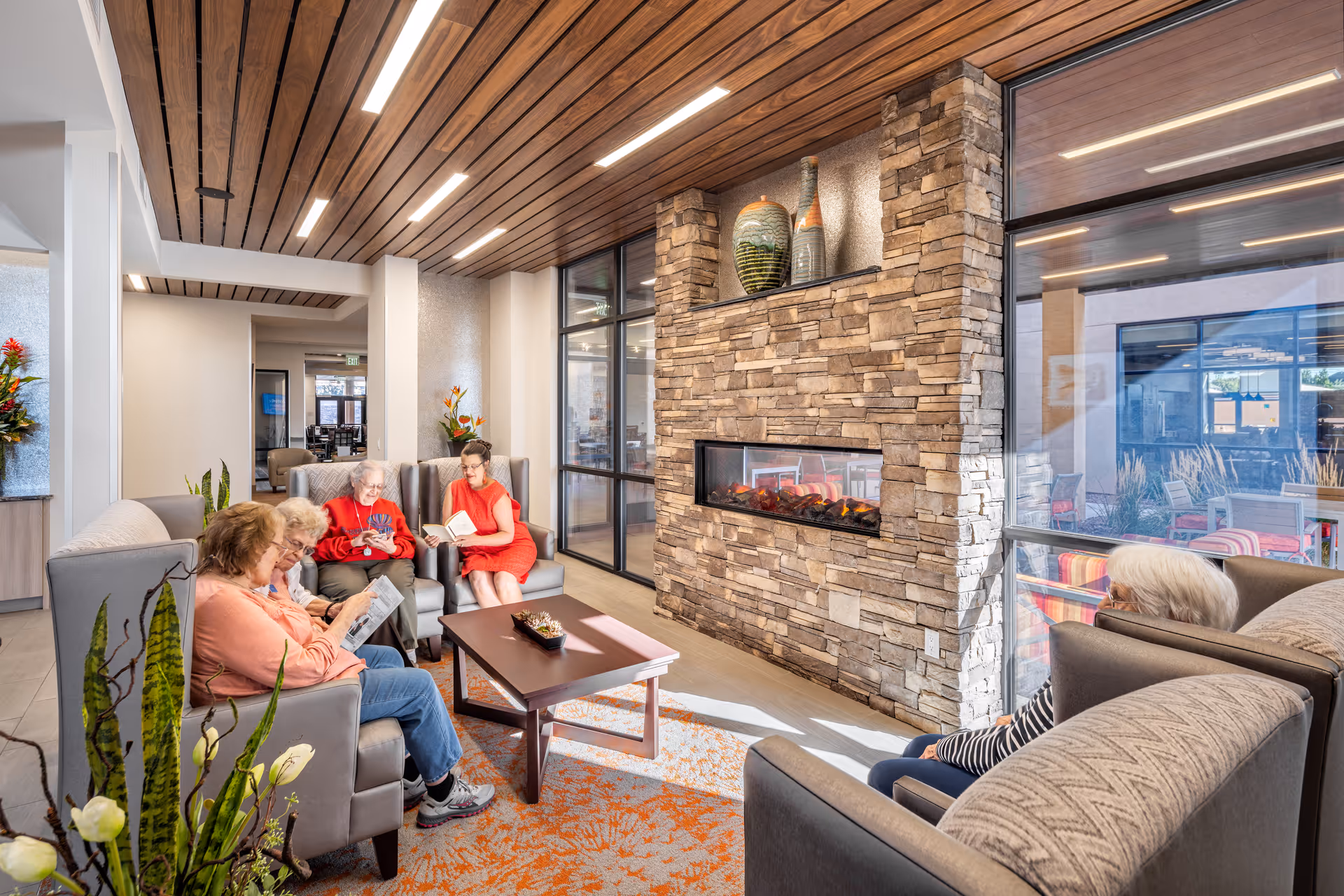 A cozy senior living common area with five elderly women sitting on comfortable armchairs around a wooden coffee table. The room features a stone fireplace with decorative vases on the mantel, large windows letting in natural light, and a wooden ceiling with recessed lighting. There are plants and flowers adding a touch of greenery to the space.