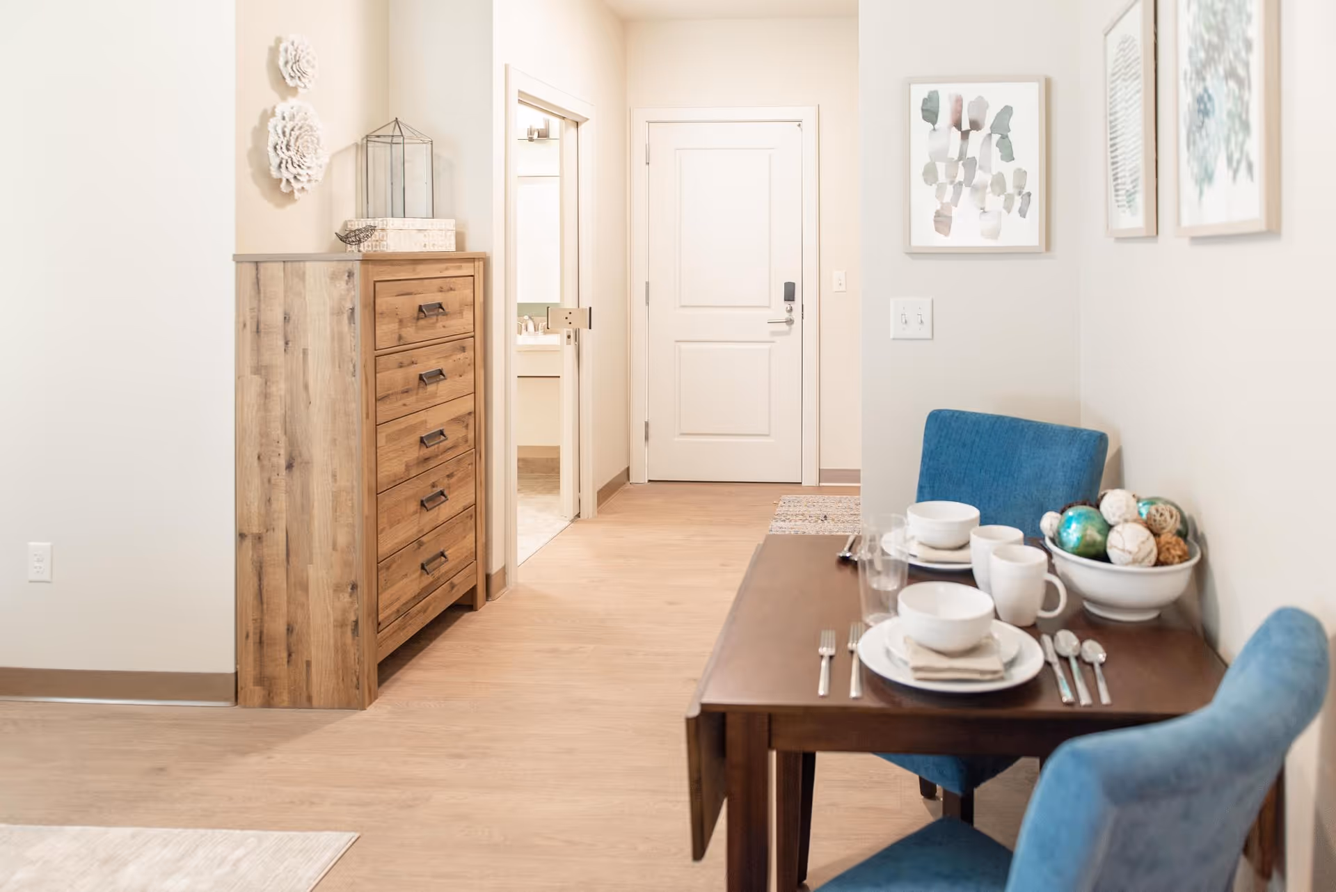 A small dining area with a wooden table set for two, featuring white dishes, cups, and silverware. Two blue upholstered chairs are positioned at the table. On the table is a decorative bowl filled with assorted decorative balls. Behind the dining area is a hallway with a closed white door and an open door leading to a bathroom. To the left of the hallway is a wooden chest of drawers with decorative items on top, and the walls are adorned with framed artwork and decorative wall hangings.