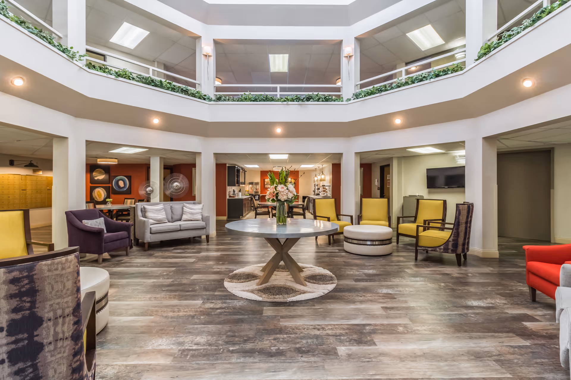 A spacious and well-lit common area in a senior living facility with a round table in the center holding a vase of flowers. Surrounding the table are various chairs in yellow, purple, and gray colors, arranged for socializing. The room has a high ceiling with an open second-floor balcony lined with greenery. The flooring is wood-style, and there are multiple doorways leading to other rooms.