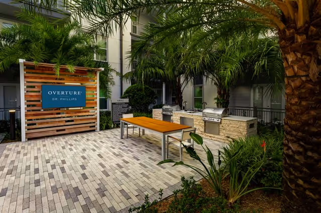 Outdoor patio area at Overture Dr. Phillips featuring a wooden table with benches, two built-in stainless steel grills, surrounded by palm trees and greenery, with a sign displaying the facility name on a wooden slat wall.